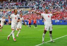 Bellingham Double Fires England Past Norway Into World Cup Semi-Finals England soccer players celebrate a goal on a green pitch with outstretched arms as fans cheer in the stadium stands behind them.