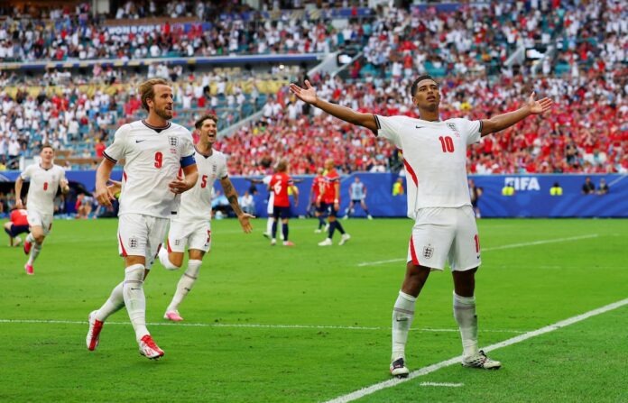 England soccer players celebrate a goal on a green pitch with outstretched arms as fans cheer in the stadium stands behind them.