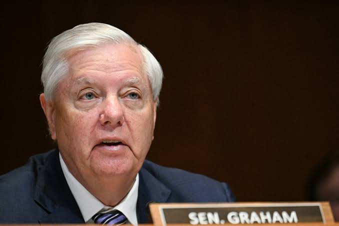 Close-up of an older man with white hair speaking at a formal hearing, wearing a dark suit and striped tie.
