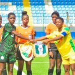 Flamingos Defeat Benin To Qualify For 2026 FIFA U-17 Women’s World Cup Women’s soccer players and referees stand on a bright green pitch as two players—one in a green-black kit and one in yellow—shake hands during pre-match rituals, with other officials in orange nearby in a stadium.