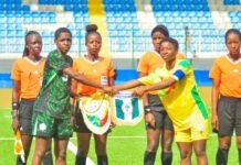 Flamingos Defeat Benin To Qualify For 2026 FIFA U-17 Women’s World Cup Women’s soccer players and referees stand on a bright green pitch as two players—one in a green-black kit and one in yellow—shake hands during pre-match rituals, with other officials in orange nearby in a stadium.