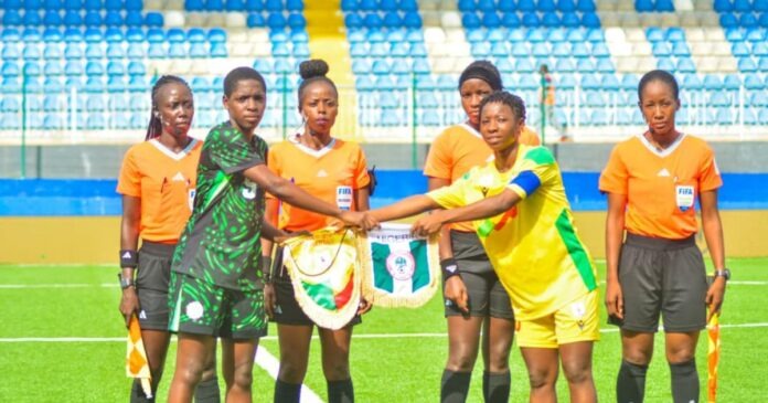 Women’s soccer players and referees stand on a bright green pitch as two players—one in a green-black kit and one in yellow—shake hands during pre-match rituals, with other officials in orange nearby in a stadium.