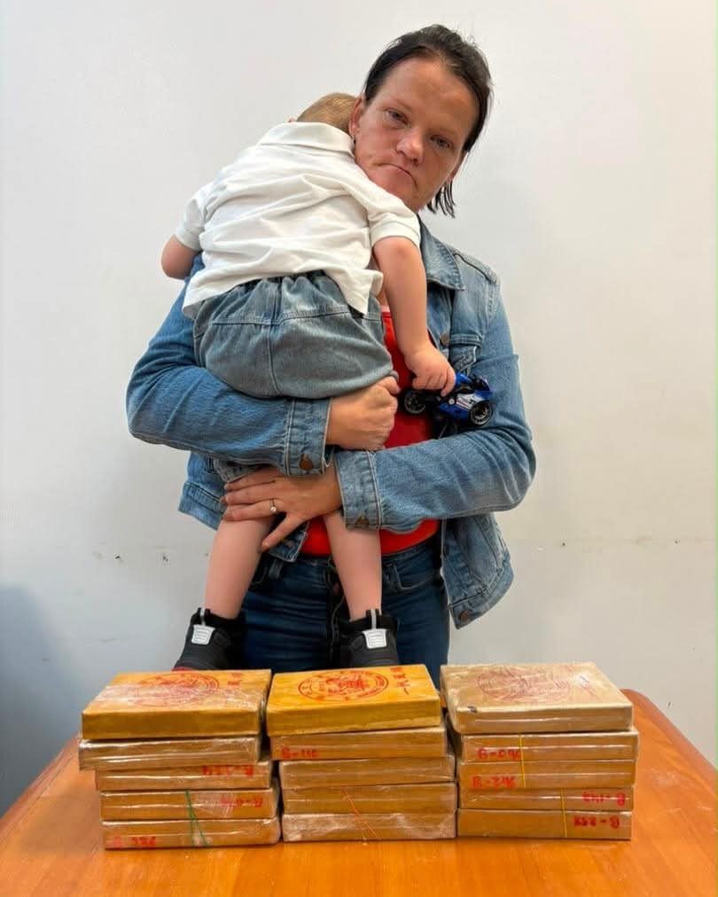 Mother in a denim jacket holds a baby while standing behind a table with stacked brown packages on it, indoors.