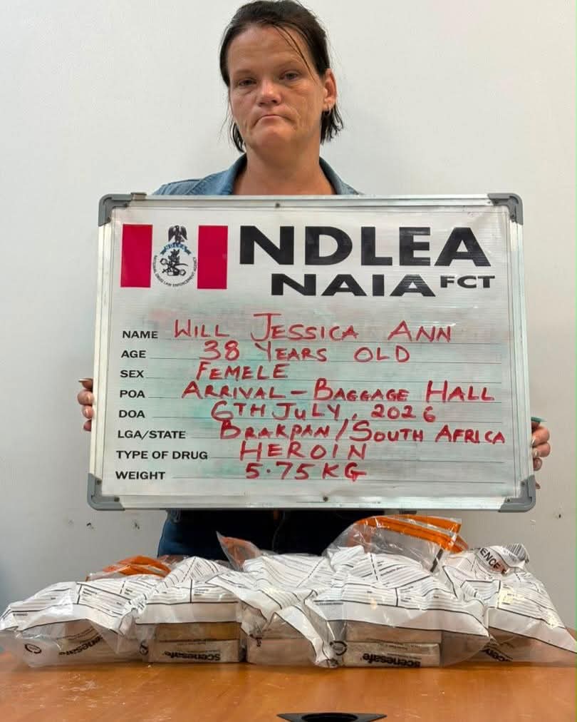 Mugshot of a woman holding an NDLEA board with handwritten details and seized packages on a table.