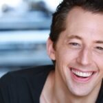 Broadway Actor Josh Grisetti Dies At 44 Close-up portrait of a smiling man with short brown hair wearing a dark shirt, facing the camera against a blurred background.