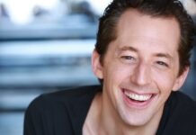 Broadway Actor Josh Grisetti Dies At 44 Close-up portrait of a smiling man with short brown hair wearing a dark shirt, facing the camera against a blurred background.