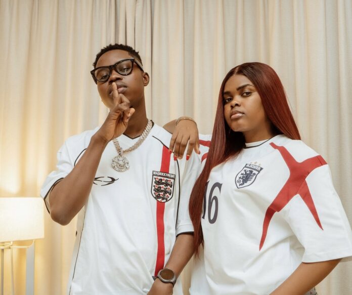 TikTok Stars Jarvis, Peller Set August 1 As Wedding Date Two people in England football jerseys pose indoors: the man with glasses makes a shushing gesture, the woman with long red hair rests her arm on his shoulder.