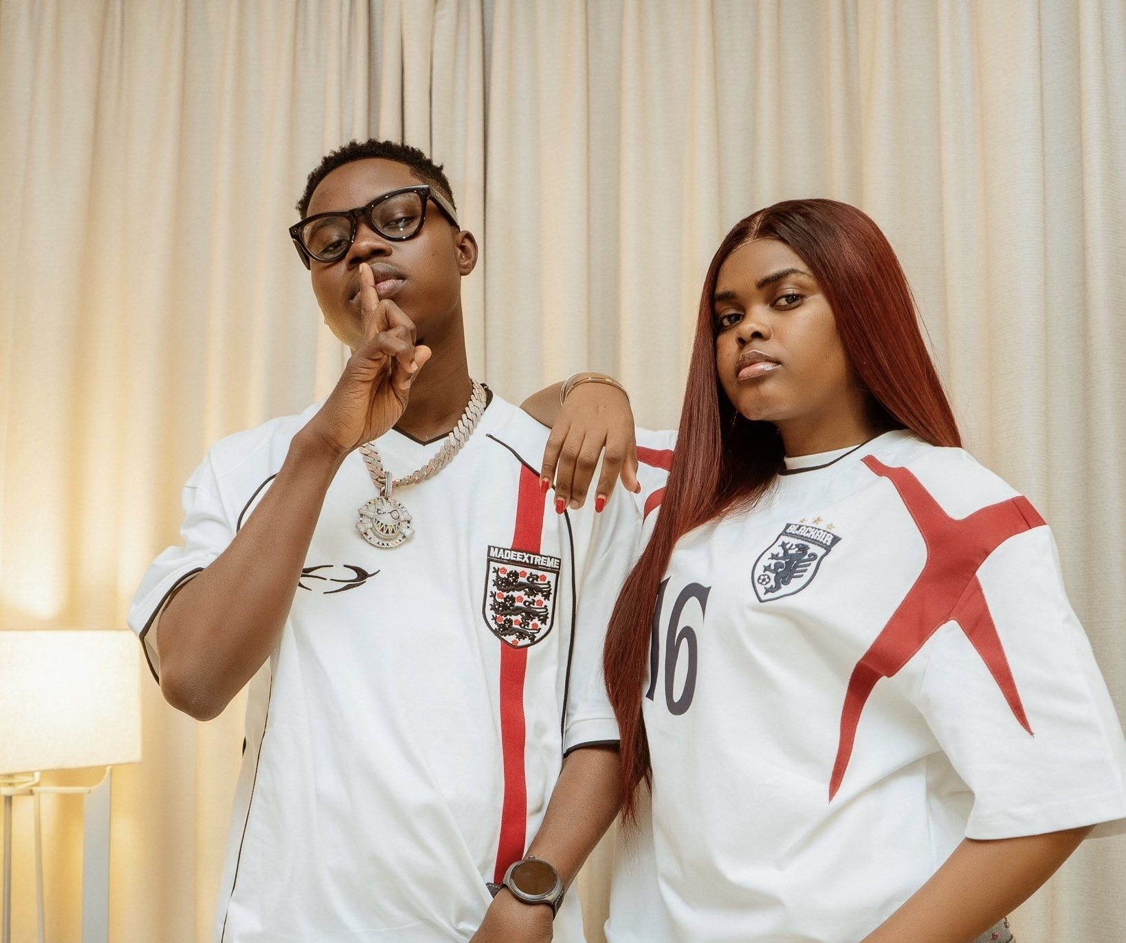 TikTok Stars Jarvis, Peller Set August 1 As Wedding Date - THEWILL NEWS MEDIA Two people in England football jerseys pose indoors: the man with glasses makes a shushing gesture, the woman with long red hair rests her arm on his shoulder.