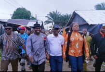 FG, International Alert Partnership Brings Eastern Obolo’s Forgotten Humanitarian Crisis Into National Focus A diverse group of people walking down a dirt street, holding hands, outdoors near wooden houses and palm trees.