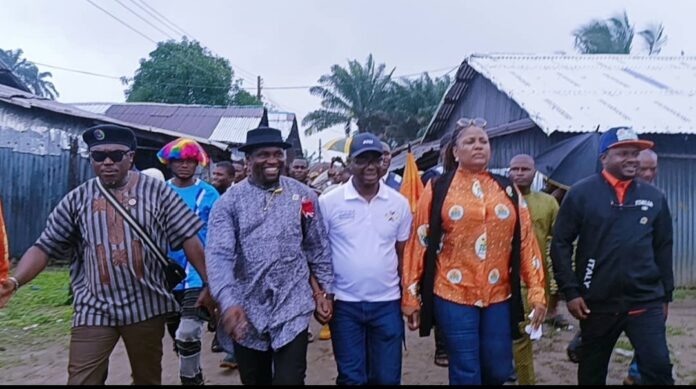 Minister's visit to communities in Eastern Obolo A diverse group of people walking down a dirt street, holding hands, outdoors near wooden houses and palm trees.