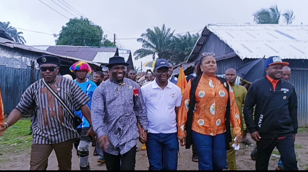 Minister's visit to communities in Eastern Obolo - THEWILL NEWS MEDIA A diverse group of people walking down a dirt street, holding hands, outdoors near wooden houses and palm trees.