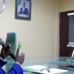 Nigeria-Hong Kong Tax Deal: 7 Ways Double Taxation Agreement Could Unlock New Opportunities For Investors And Traders Man in a blue suit speaks into a handheld microphone during a virtual meeting, with a Nigerian flag on the desk and a video call on a large screen.