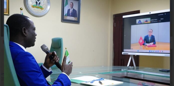 Man in a blue suit speaks into a handheld microphone during a virtual meeting, with a Nigerian flag on the desk and a video call on a large screen.
