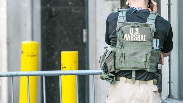 Deputy U.S. Marshal Back view of a U.S. Marshal wearing a green tactical vest and tan cargo pants, talking on a radio.