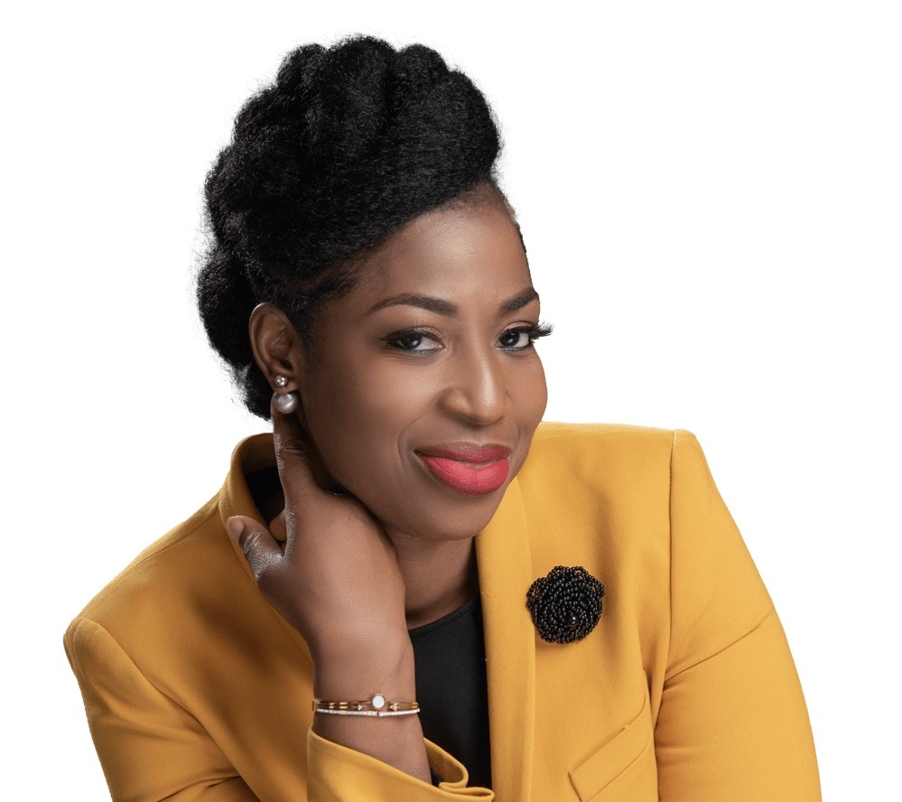 Portrait of a smiling Black woman with natural hair in a mustard blazer, posing with her hand at her chin and wearing a black top and brooch accessory.