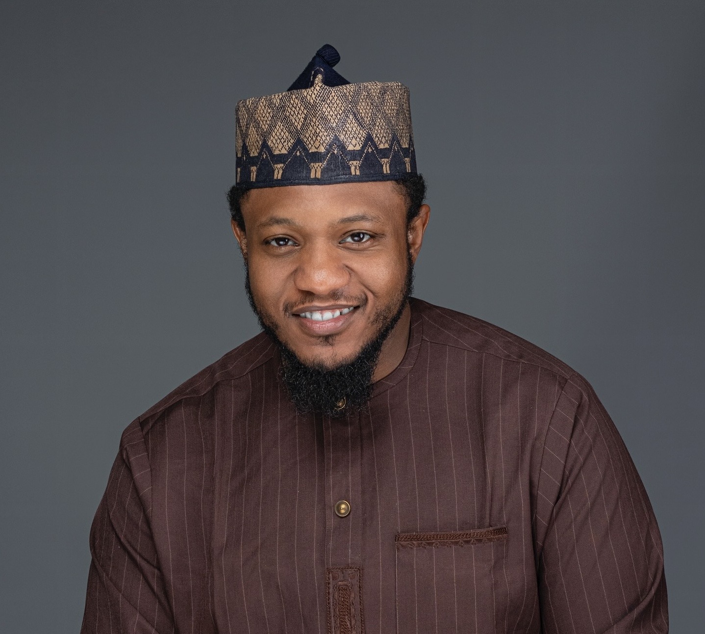 Smiling man wearing a patterned traditional cap and brown striped shirt against a grey background