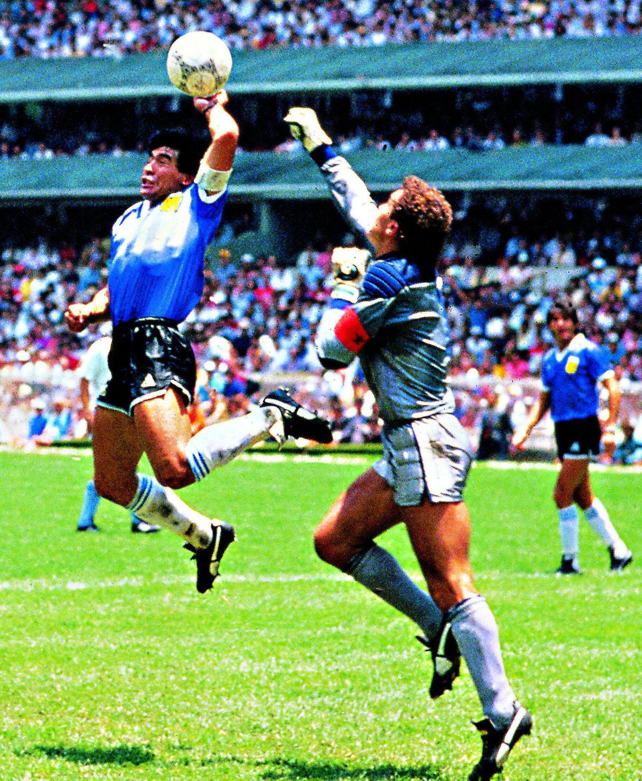 Argentine footballer Diego Maradona (left) scoring with his hand past English goalkeeper Peter Shilton. - THEWILL NEWS MEDIA Two soccer players jump for a high ball during a match, one in a blue jersey and black shorts reaching upward while the goalie in gray with gloves defends nearby as a crowd watches from the stands.