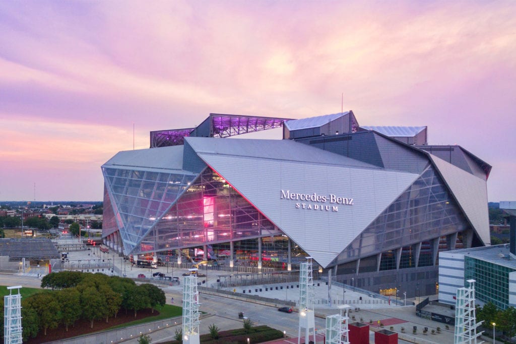Mercedes-Benz Stadium - THEWILL NEWS MEDIA Mercedes-Benz Stadium illuminated at sunset with pink and purple sky behind the angular glass exterior