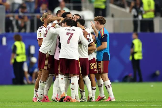 Soccer players in white and maroon jerseys huddle together on the green pitch, Ferran Torres wearing number 7 at the center.