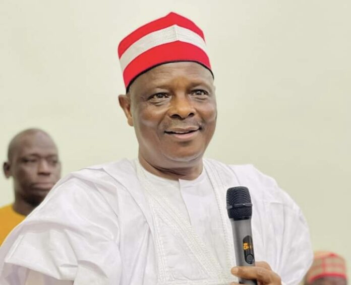 NDC Vice Presidential candidate, Rabiu Kwankwaso. Man in white traditional attire holding a microphone, speaking at an event, red and white cap on his head.",