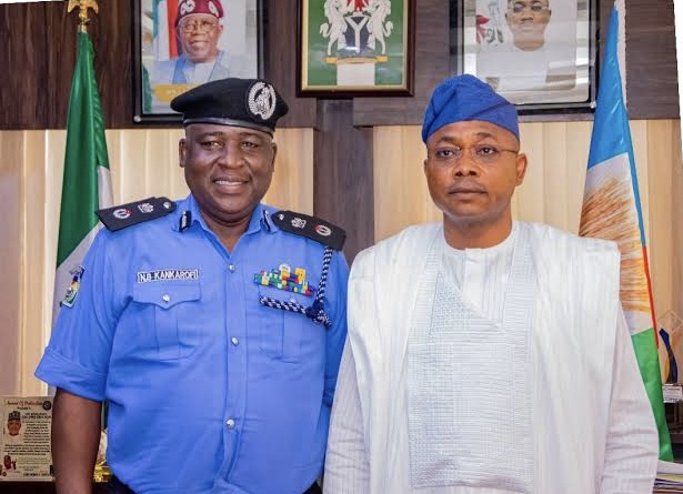 From L: CP Naziru Bello Kankarofi and Governor Ahmed Usman Ododo Two men pose in an office: a Nigerian police officer in blue uniform and a man in white traditional attire with a blue cap, with flags in the background.