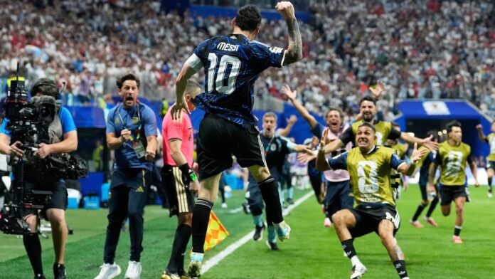 Messi (number 10) leaps in celebration on a soccer field as photographers and teammates run toward him and a packed stadium cheers behind them.