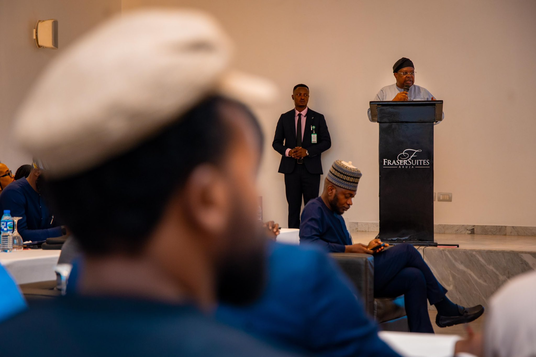 Minister of Power, Joseph Tegbe - THEWILL NEWS MEDIA Speaker at a podium delivering a talk in a conference room; the Fraser Suites Abuja logo is visible on the lectern. Attendees are seated in the foreground, out of focus.