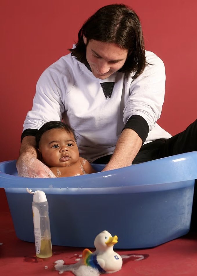 An iconic UNICEF charity photograph taken in 2007 shows a young Lionel Messi bathing six-month-old Lamine Yamal Photo: Joan Monfort/UNICEF/Sport. - THEWILL NEWS MEDIA Adult caregiver bathes a baby in a blue plastic tub against a red background, with a rubber duck toy nearby.