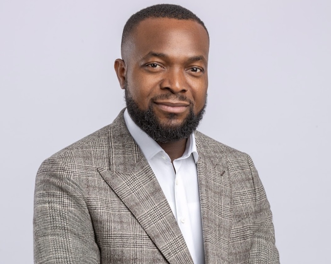 Professional headshot of a smiling man in a gray plaid blazer and white shirt, neutral background.
