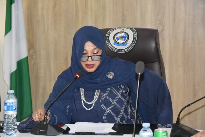 Intelligence Unit (NFIU). Woman in a navy hijab and glasses speaks into a microphone at a formal meeting, with documents on the table and a seal on the chair behind her.