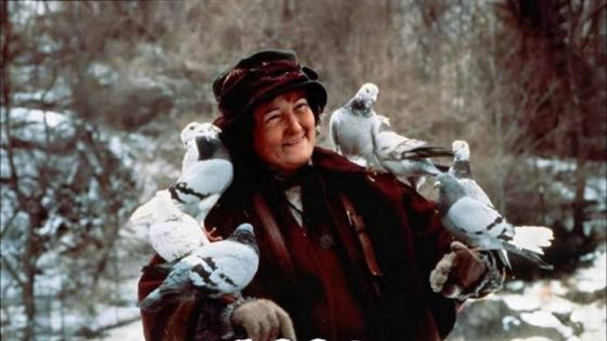 Brenda Fricker, the kind-hearted Pigeon Lady in Home Alone 2. Credit: 20th Century Fox/Kobal/Shutterstock - THEWILL NEWS MEDIA Smiling woman in a dark red winter coat with multiple white pigeons perched on her gloved arms in a snowy forest setting.