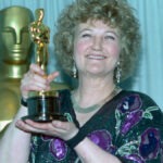 Brenda Fricker, Oscar Winner and ‘Home Alone 2’ Star, Dies at 81 Smiling woman with curly hair holding an Oscar trophy in front of a gold statuette backdrop.