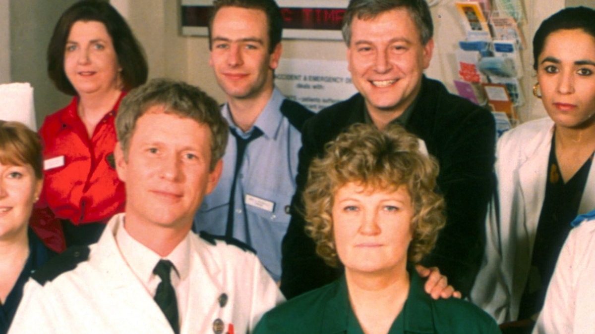Brenda Fricker (center) as Megan Roach in Casualty. (Image credit: BBC) - THEWILL NEWS MEDIA Group of healthcare workers posing for a photo in a clinic, with a man in a white coat at the front.