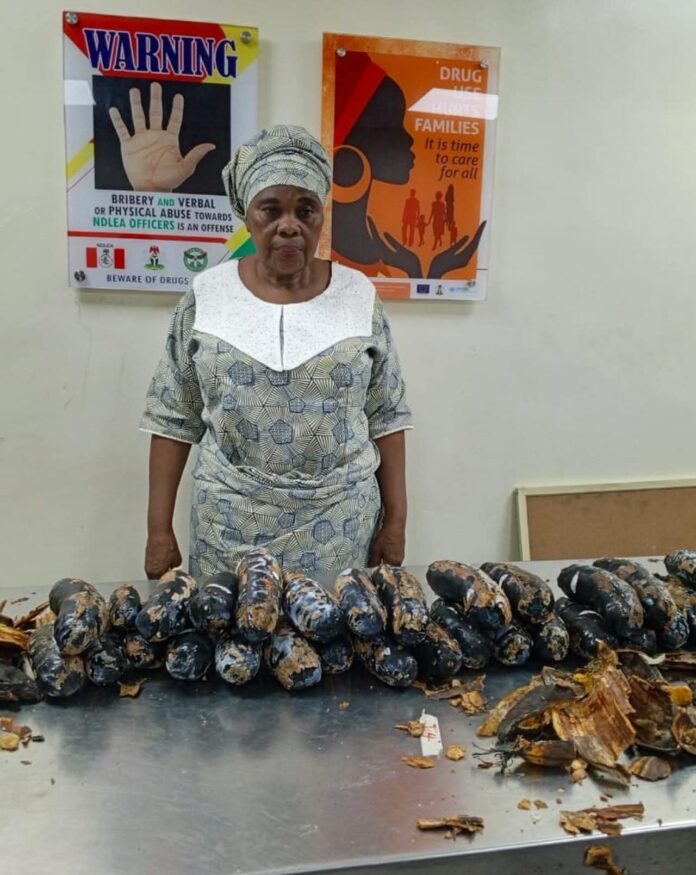 Elderly woman wearing a patterned dress and headwrap stands behind a table filled with rows of dark, cylindrical food items wrapped in leaves/peels on a metal surface.