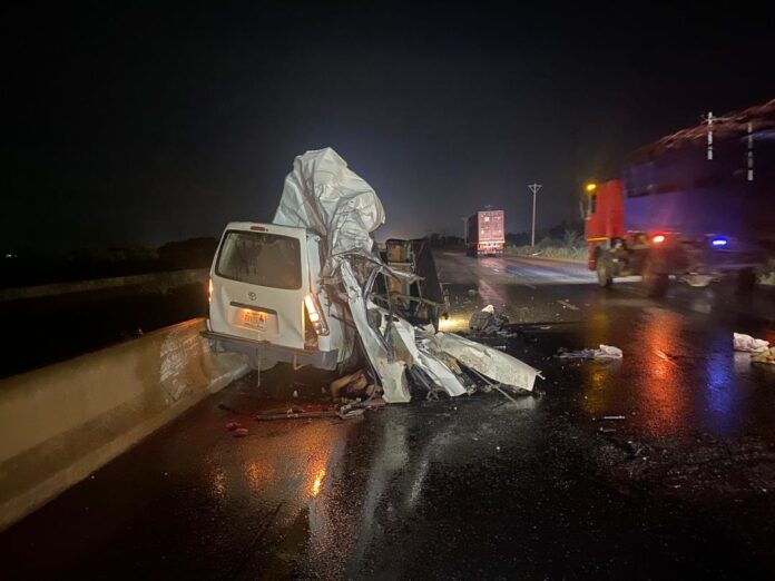 IMG_6649 White van severely damaged on a rainy highway at night, debris scattered and emergency lights in the distance.