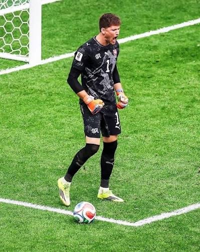 Gregor Kobel Soccer goalkeeper in black kit with orange gloves standing near a goal, ball at his feet on the green pitch.