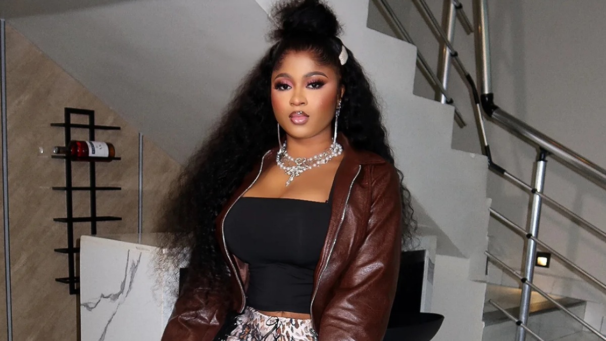 Imisi - THEWILL NEWS MEDIA Woman with long curly hair wearing a brown leather jacket over a black strapless top, standing indoors by a staircase.