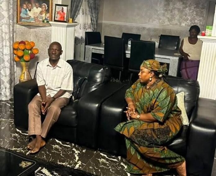 Two adults sit on black leather couches in a living room, facing each other and chatting, with a vase of orange flowers and framed photos in the background.