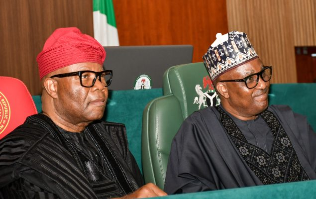 NASS Two Nigerian men in traditional attire sit side by side at a formal conference table, with a flag and green chairs in the background.