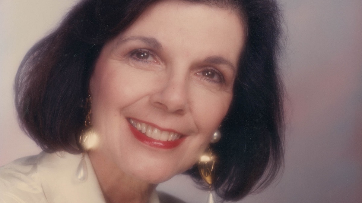 Mary Jo 2 - THEWILL NEWS MEDIA Close-up portrait of a smiling woman with dark hair, red lipstick, and pearl earrings in soft lighting.