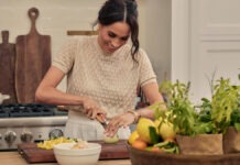 Duchess Of Sussex Meghan Markle Scores First Daytime Emmy Nomination For Netflix Series ‘With Love, Meghan’ Woman chopping an onion on a wooden cutting board in a bright kitchen, with herbs and a basket of fruit nearby