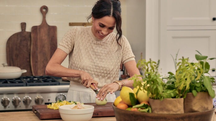 Meghan Markle, Duchess of Sussex. Credit: Chris Jackson/Getty Images | ‘With Love, Meghan’ Netflix poster. Credit: Meghan via Instagram Woman chopping an onion on a wooden cutting board in a bright kitchen, with herbs and a basket of fruit nearby