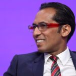 Tanzania’s Richest Man Mohammed Dewji to Invest $275 Million in Graphite Mining, Expand Luxury Tourism Man in a navy suit and red-framed glasses smiles during a talk on a purple stage backdrop with a mic nearby.