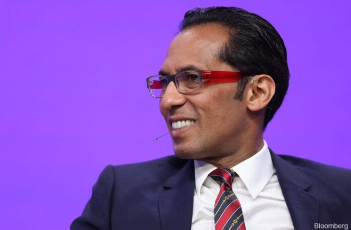 Mohammed_Dewji Man in a navy suit and red-framed glasses smiles during a talk on a purple stage backdrop with a mic nearby.