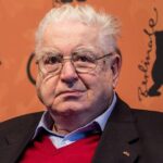 Legendary Film Festival Director Moritz De Hadeln Dies At 85 Older man with white hair and glasses wearing a red sweater and dark blazer, posed against an orange backdrop with logos.