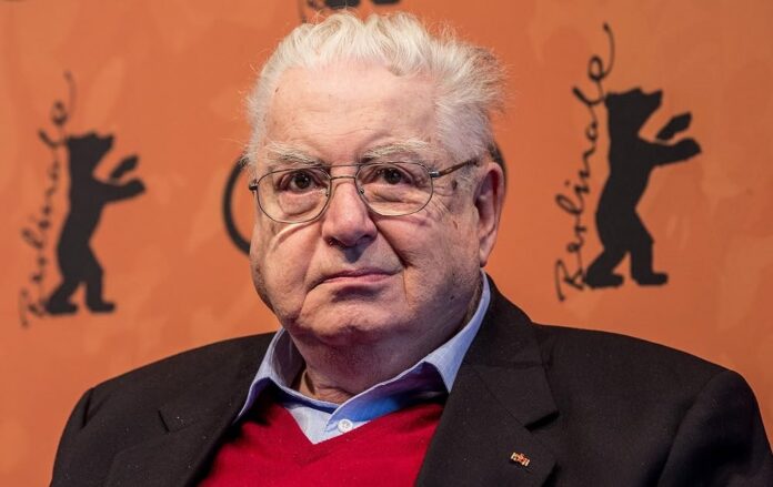 Moritz De Hadeln Older man with white hair and glasses wearing a red sweater and dark blazer, posed against an orange backdrop with logos.