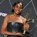 Nigerian-American Actresses Ayo Edebiri, Joy Sunday Earn Emmy Nominations as ‘The Pitt’ Leads With 25 Nods Smiling woman in a strapless black dress holding an Emmy Award on the red carpet with a patterned backdrop behind her.