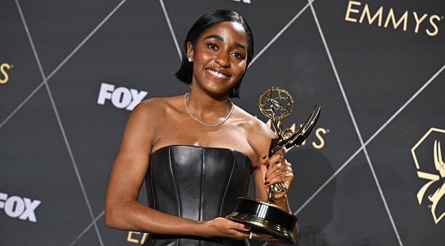 Smiling woman in a strapless black dress holding an Emmy Award on the red carpet with a patterned backdrop behind her.