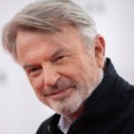 Jurassic Park Star Sam Neill’s Cause Of Death Revealed After Beating Cancer Portrait of a smiling older man with gray hair and a light beard, wearing a dark shirt.