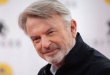Jurassic Park Star Sam Neill’s Cause Of Death Revealed After Beating Cancer Portrait of a smiling older man with gray hair and a light beard, wearing a dark shirt.